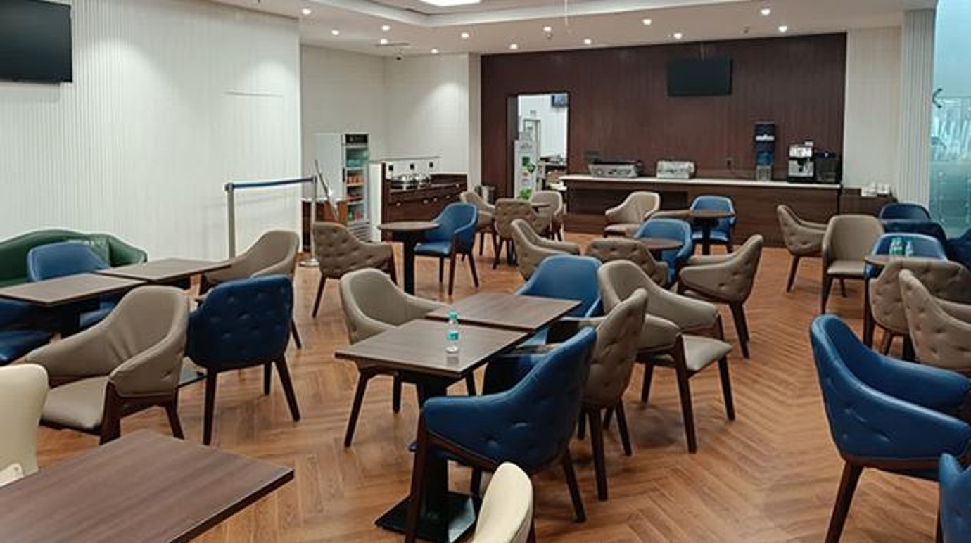 The Lounge at Ahmedabad Airport (AMD) Terminal 2 featuring modern seating with blue and beige chairs, wooden tables, and a staffed food and beverage counter in the background. The Lounge at Ahmedabad Airport (AMD) Terminal 2 featuring modern seating with blue and beige chairs, wooden tables, and a staffed food and beverage counter in the background.