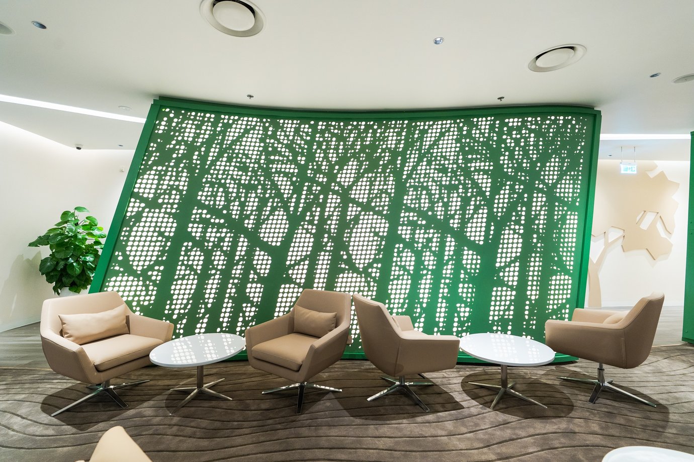 Modern seating area inside Vanda Lounge at Da Nang International Airport, featuring beige armchairs, circular coffee tables, and a green perforated wall panel inspired by tree patterns for a tranquil atmosphere.