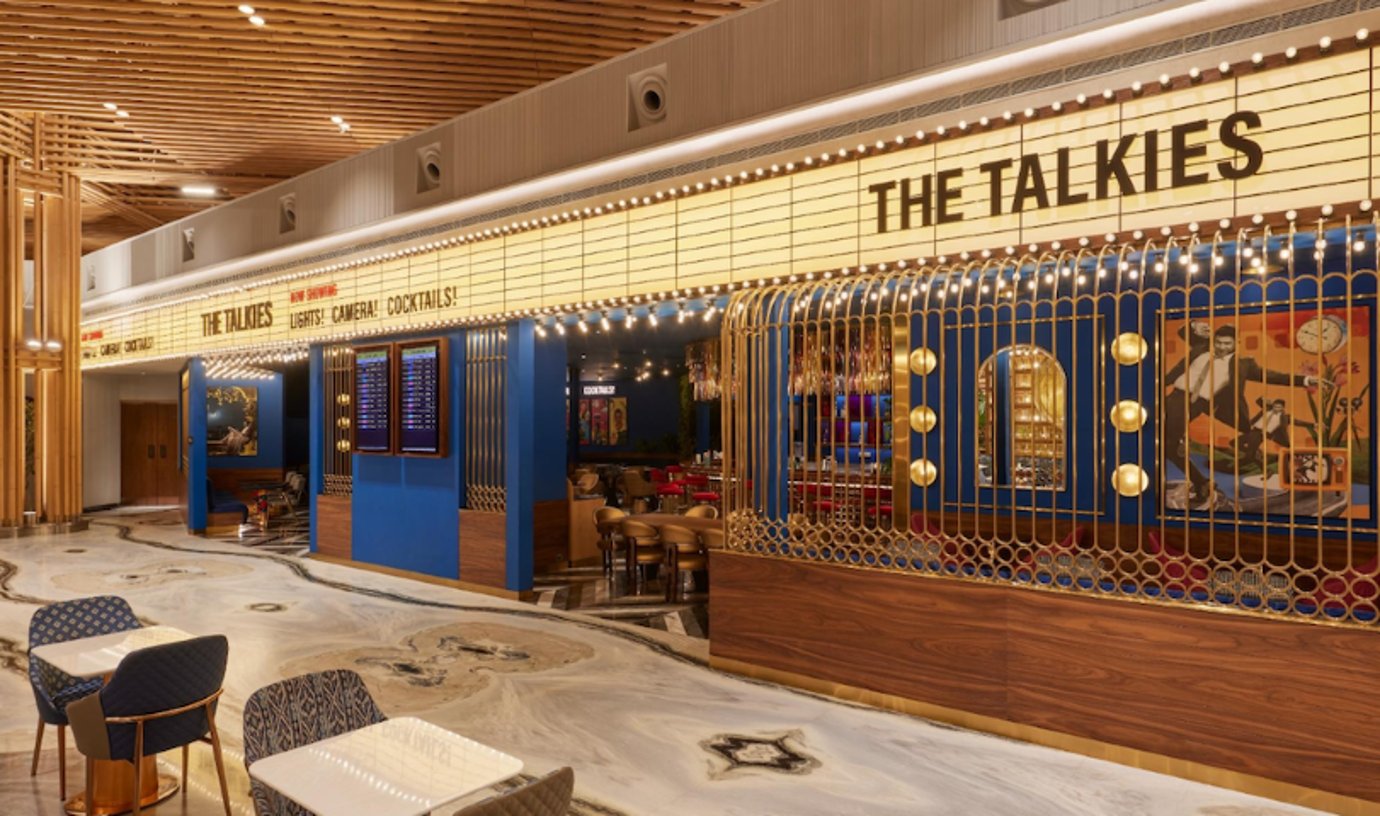 Exterior view of 'The Talkies' lounge area at Terminal 2, Bangalore Airport, designed to resemble a vintage movie theater. The facade features a classic marquee with illuminated letters reading 'The Talkies'.