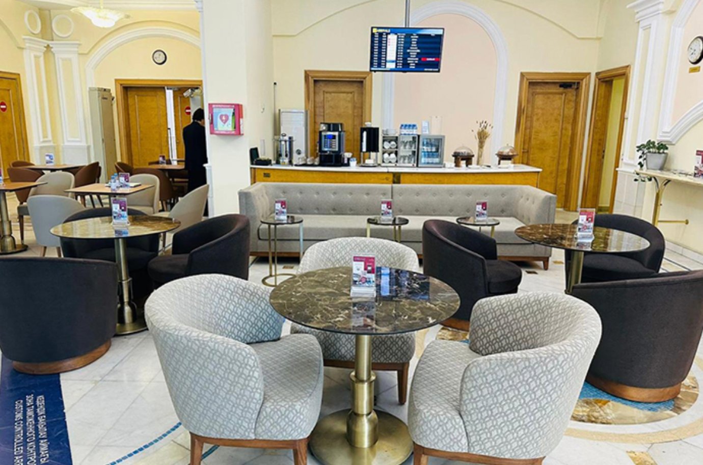Cozy seating area inside the Primeclass Lounge at Almaty International Airport, featuring round marble tables, soft grey and brown armchairs, and a refreshment counter with coffee machines and snacks.