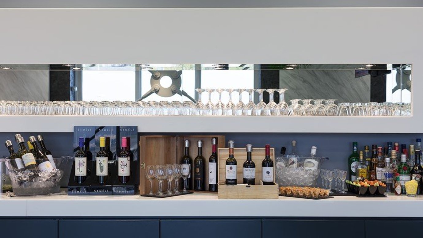 Beverage station at the Goldair CIP lounge at Athens Airport. The station displays mainly a variety of alcoholic beverages, mostly red and white wine, some in ice buckets. To the side is neatly arranged spirits and liquor. On the top shelf is glasses