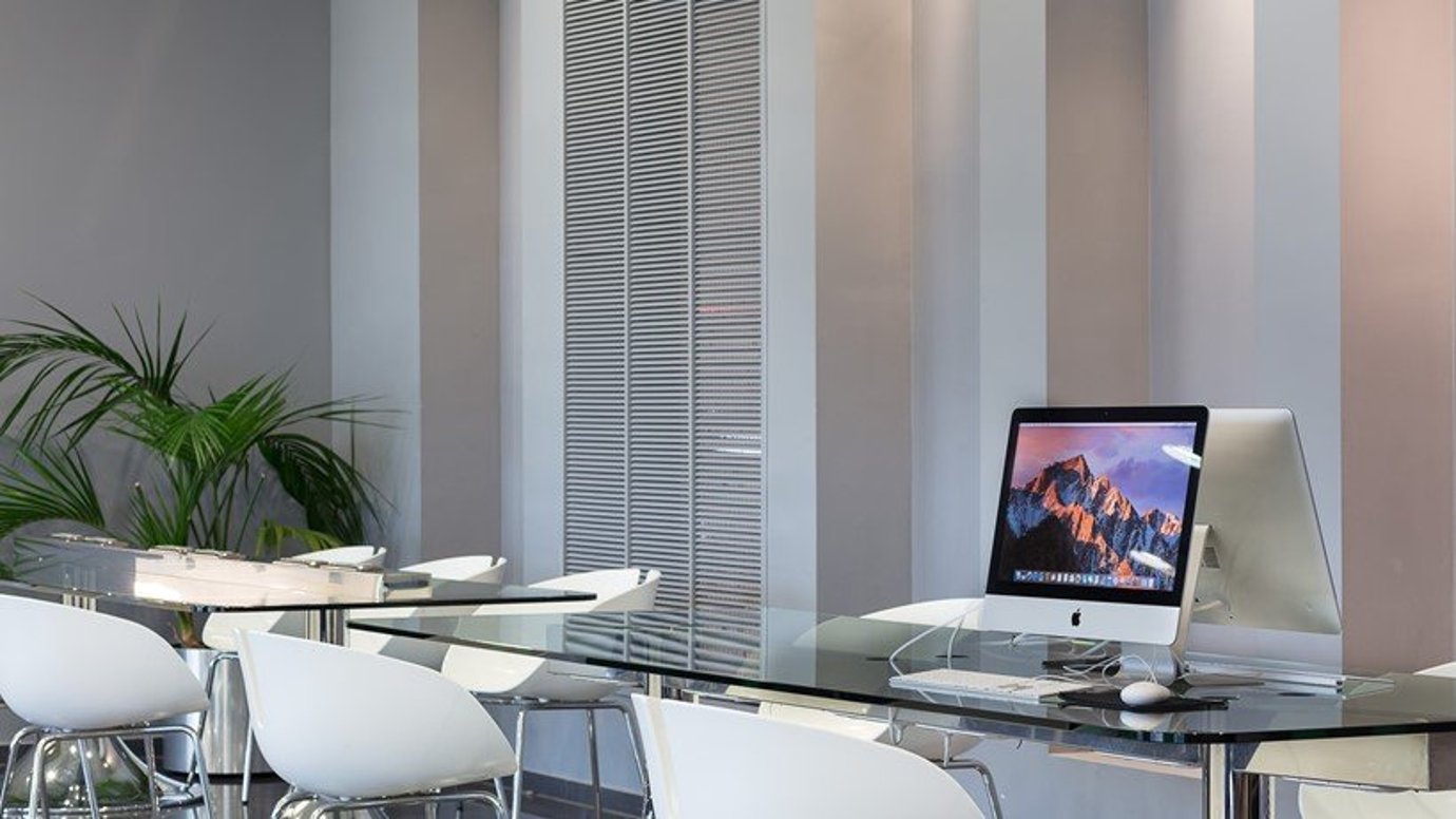 Modern workspace area in the goldair CIP lounge at Athens Airport, featuring sleek glass top tables and ergonomic white chairs. There are two large iMacs on one of the tables displaying mountain desktop wallpaper. A plant rests in the background.