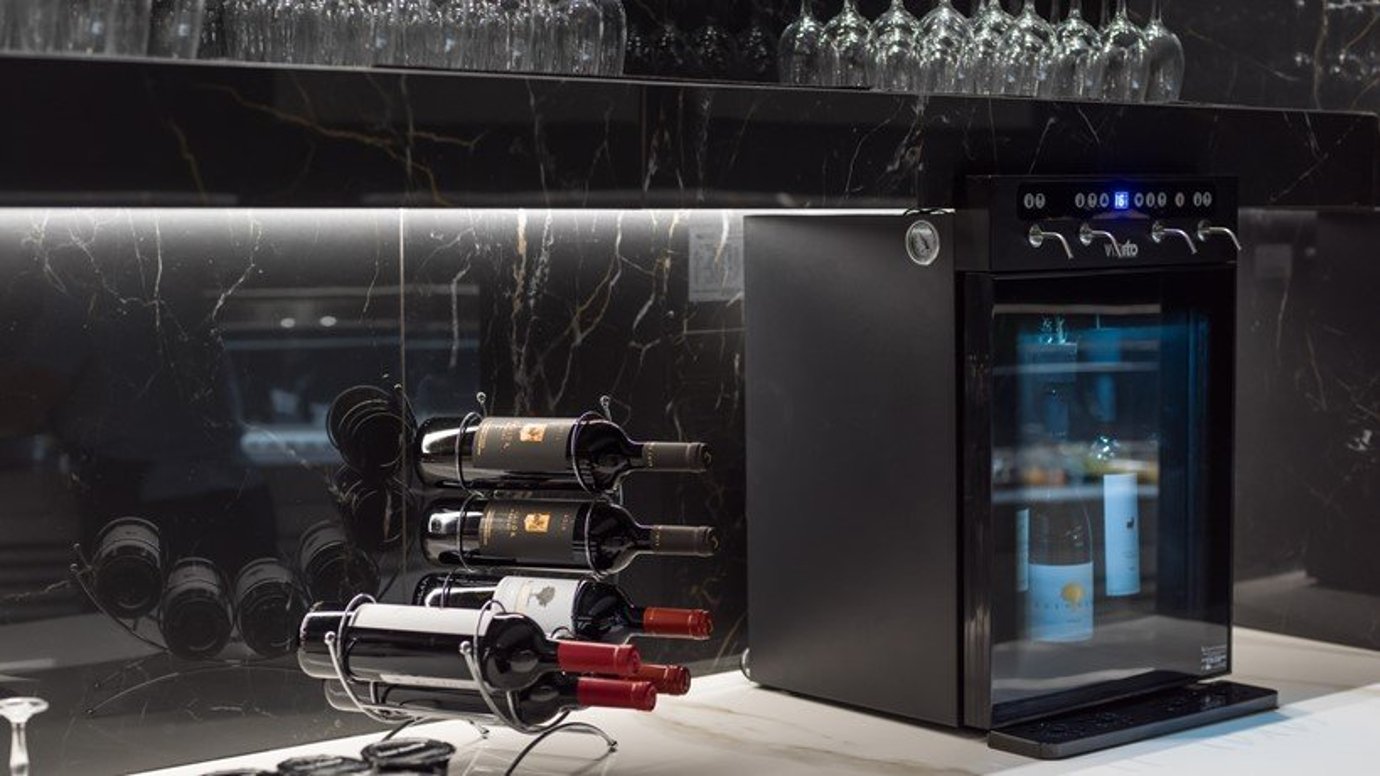Close-up of a sophisticated wine storage area at the Goldair Handling Lounge at Athens Airport, showcasing a sleek black wine fridge with wine inside. Adjacent to the fridge is a rack holding several bottles of red wine.