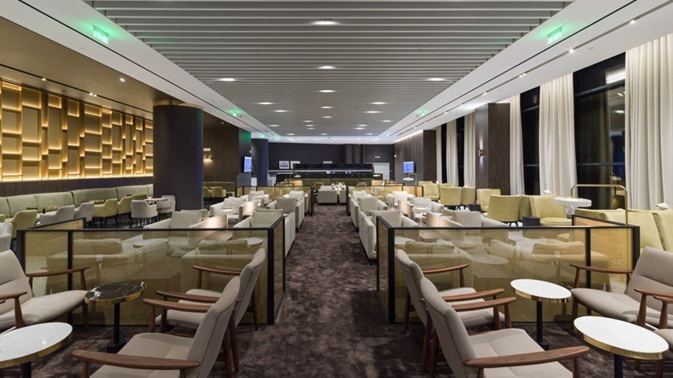 Spacious seating area in the Goldair Handling Lounge, featuring an array of cream coloured armchairs and sofas with modern glass topped coffee tables. The lounge is lit by soft overhead lighting and decorated with green ambient lighting accents.