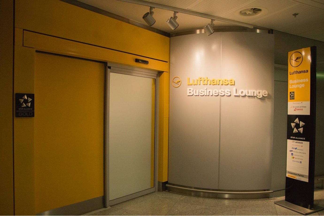 Entrance area to the Lufthansa Business Lounge at Athens Airport, featuring the company's logo and large signage. A sign post is to the side of the door detailing entry requirements to the lounge.