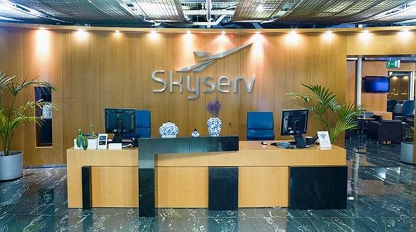 Skyserv Aristotle Onassis reception desk at Athens International Airport. The skyserv singage hangs against a wooden back wall, coupled with a stylised wooden desk. Decorative flora is situated on either side of the desk, as well as on top of it.