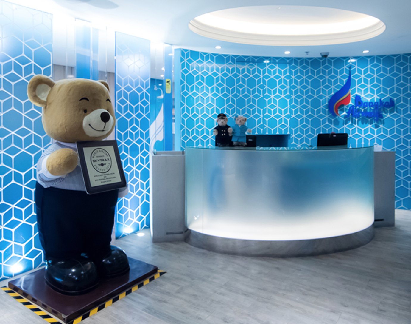 Bangkok Airways Boutique Lounge reception at Suvarnabhumi Airport featuring signature blue geometric walls, airline mascot bear, and frosted glass check-in counter.