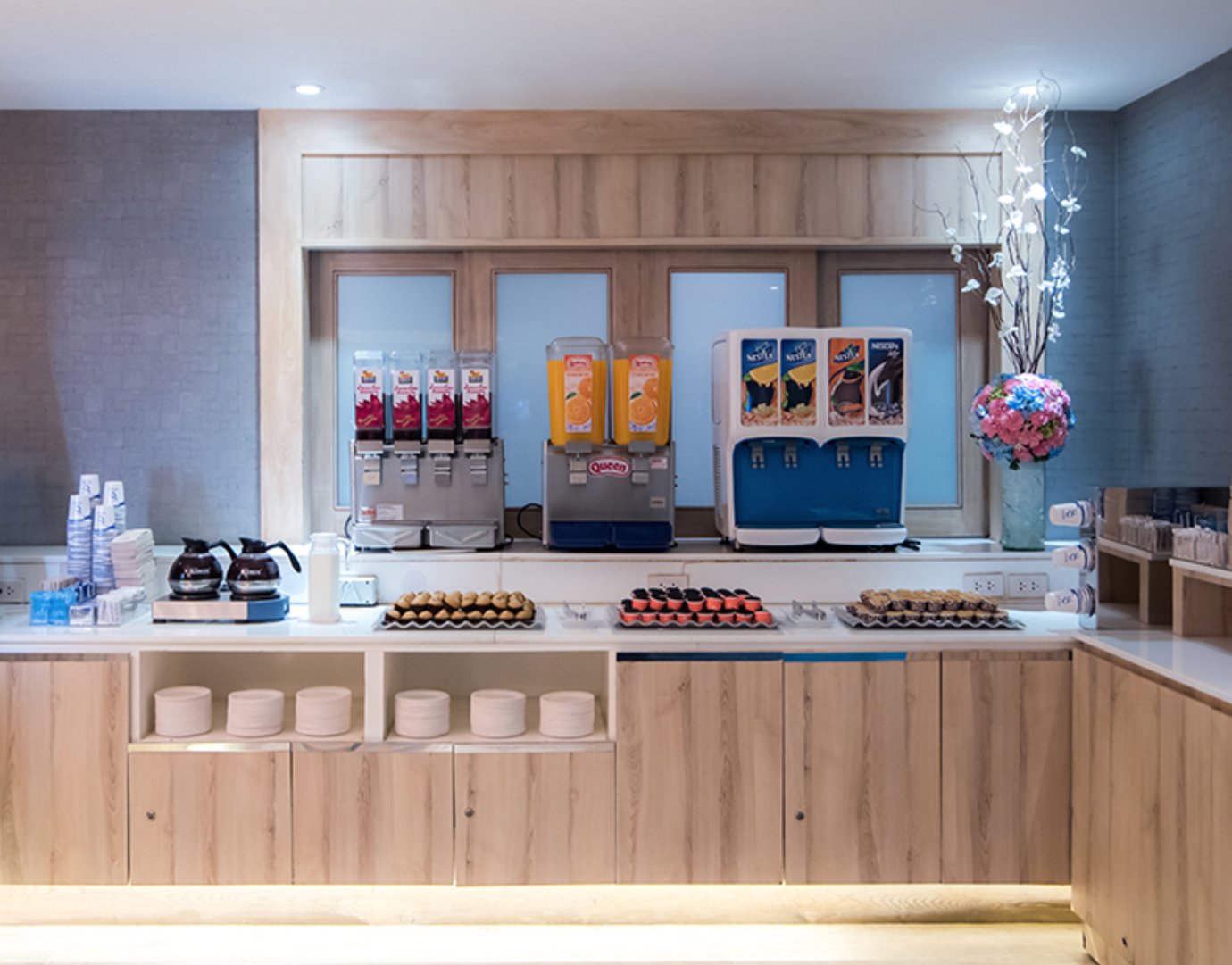 Beverage and snack station at the Bangkok Airways Boutique Lounge in Suvarnabhumi Airport, featuring juice dispensers, coffee pots, and light wood counters.