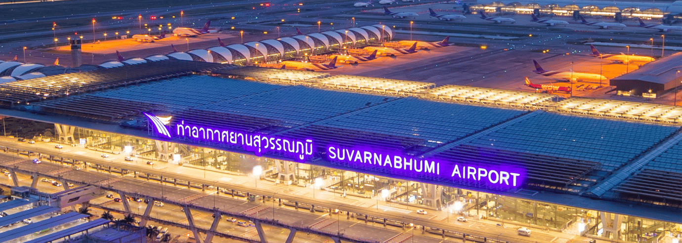 Aerial evening view of Suvarnabhumi Airport in Bangkok, Thailand, showing the illuminated terminal building, parked aircraft, and runway lights.