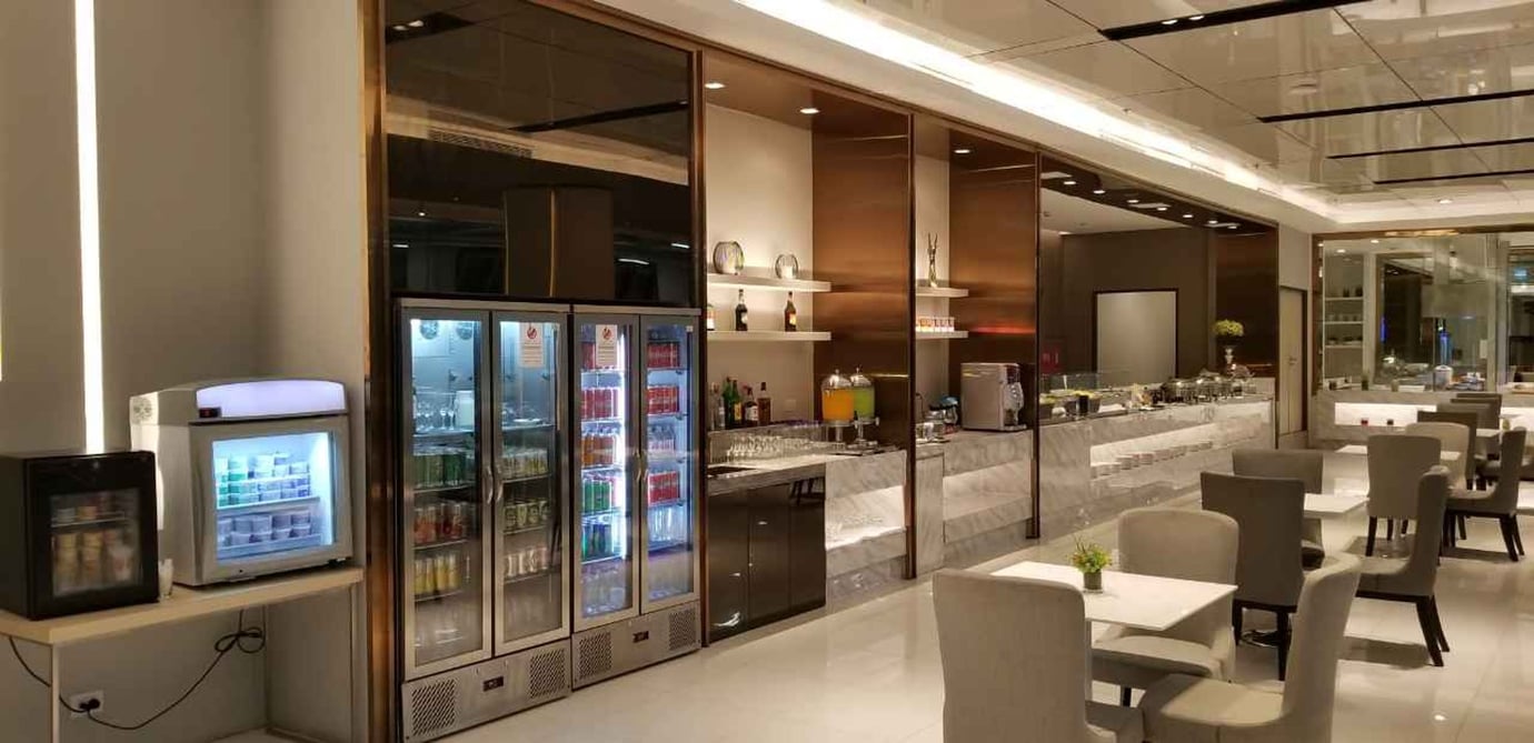 Buffet and beverage counter at the Miracle First Class Lounge Concourse D Bangkok Suvarnabhumi Airport with refrigerators stocked with soft drinks, juices, and snacks, plus a marble service bar.
