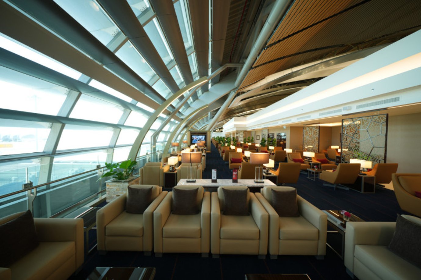 Interior of Emirates Lounge Satellite 1 Bangkok Suvarnabhumi Airport showing rows of beige leather armchairs, soft lamps, and large curved windows overlooking the tarmac.