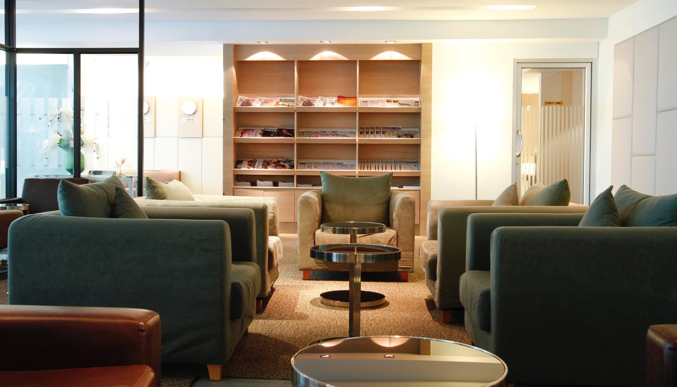 Seating area inside the Miracle First and Business Class Lounge at Bangkok Suvarnabhumi Airport featuring soft green and beige armchairs arranged around low circular tables, carpeted flooring, and a wooden bookshelf filled with magazines and newspapers