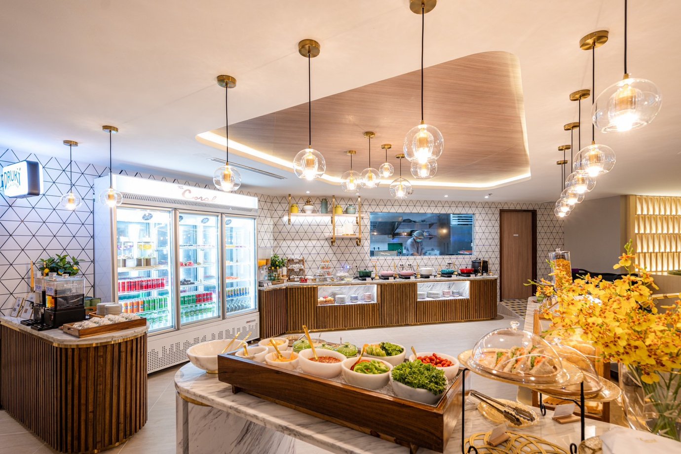 The Coral Finest Business Class Lounge buffet area at Bangkok Suvarnabhumi Airport featuring marble countertops with fresh salads, sandwiches, and desserts, and a backlit refrigerator filled with drinks