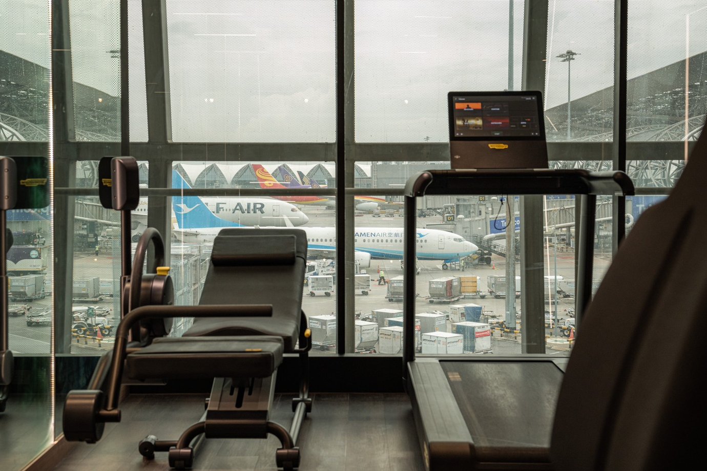 Fitness room at The Coral Finest Business Class Lounge, Bangkok Suvarnabhumi Airport, featuring a treadmill and workout bench positioned beside large windows