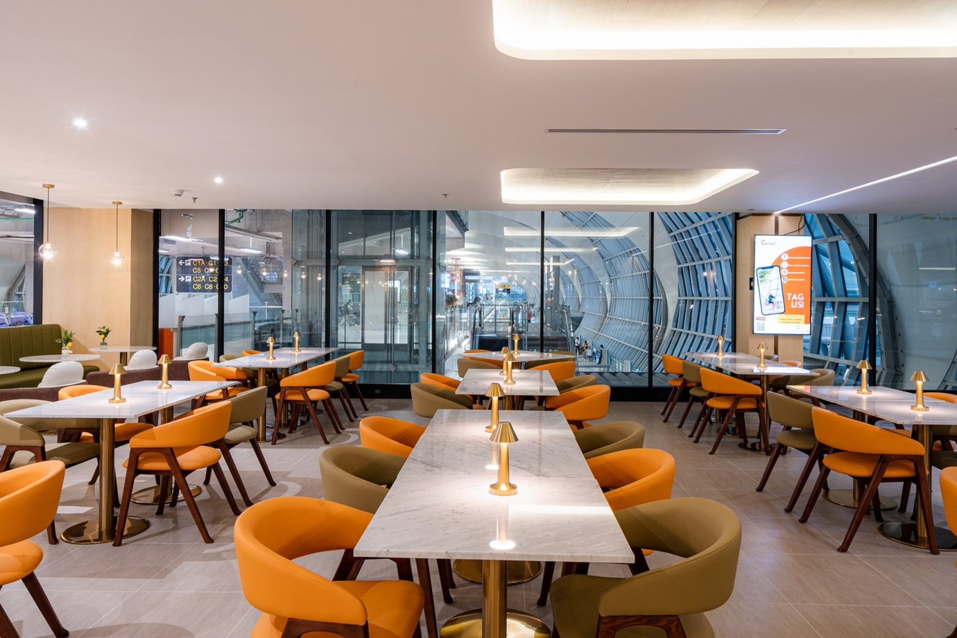 Dining area inside The Coral Finest Business Class Lounge at Bangkok Suvarnabhumi Airport featuring long marble tables, gold accent lamps, and modern chairs in orange and beige tones, set against floor-to-ceiling glass windows