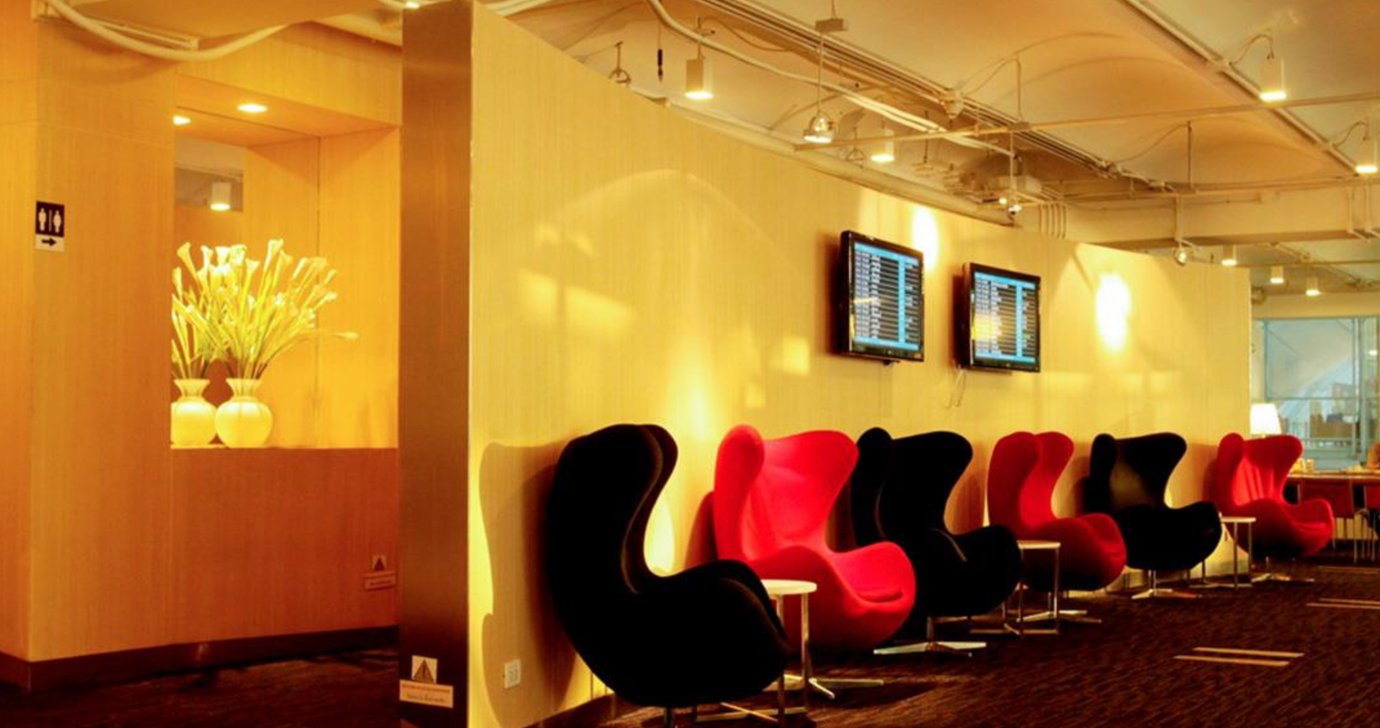 Seating area at Miracle First Class Lounge Concourse G Bangkok Suvarnabhumi Airport featuring red and black modern chairs, soft lighting, and flight information screens on the wall.