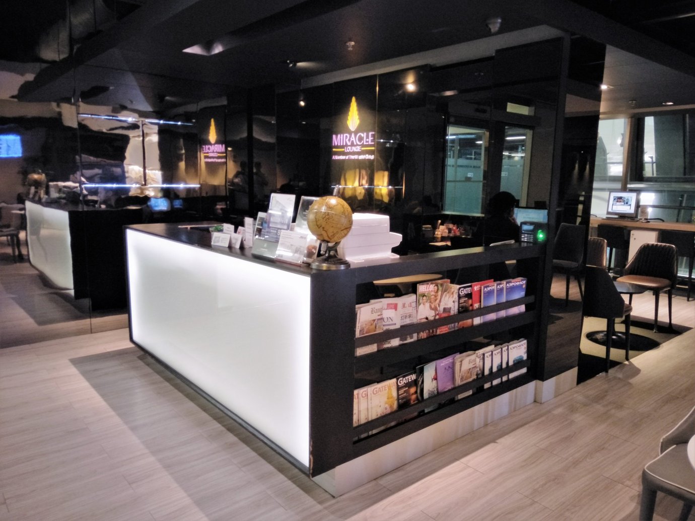 Reception counter inside the Miracle Lounge, Concourse C, Bangkok Suvarnabhumi Airport, featuring a sleek illuminated desk with travel brochures and magazines on display, dark reflective walls, and a seating area visible in the background.