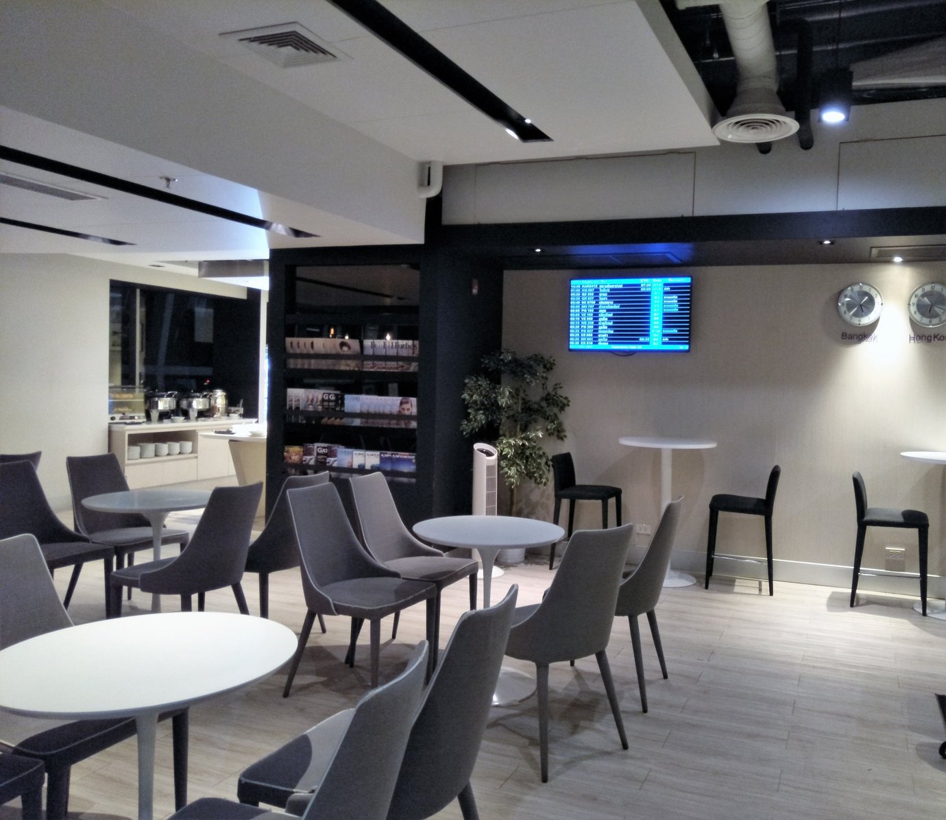 Main seating area inside the Miracle First Class Lounge, Concourse C, Bangkok Suvarnabhumi Airport, featuring rows of grey upholstered chairs and white circular tables, a buffet counter in the background, and a wall-mounted flight information screen.