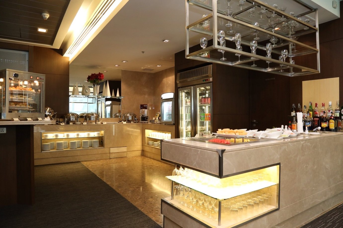 Buffet and bar counter inside the Miracle First and Business Class Lounge, Concourse A, Bangkok Suvarnabhumi Airport, featuring marble surfaces, glass shelves with wine glasses, beverage fridges stocked with drinks, and a selection of food and pastries on display.