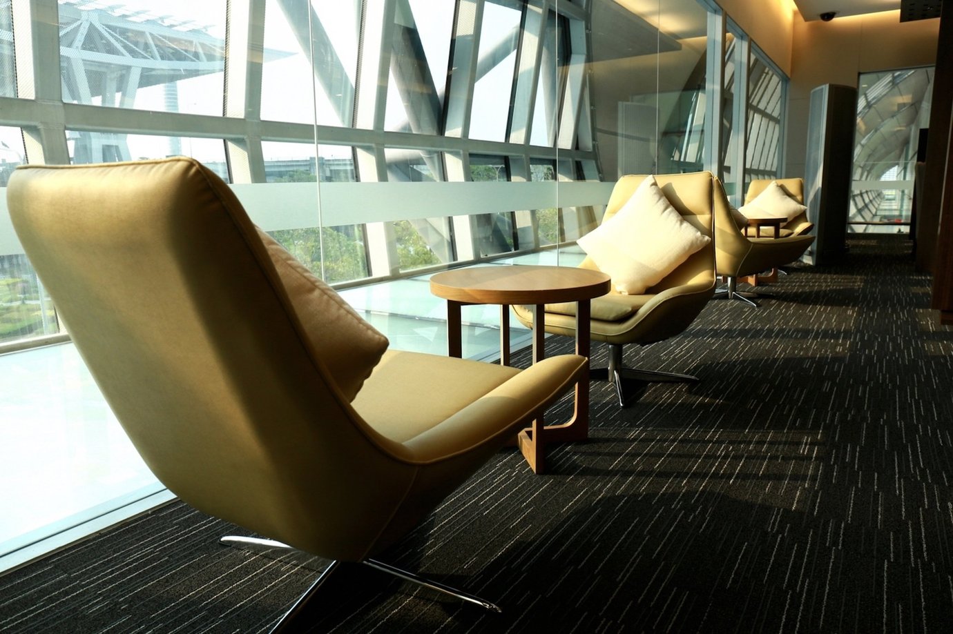 Row of tan leather lounge chairs with soft cream cushions positioned beside a floor-to-ceiling glass window at the Miracle First and Business Class Lounge, Concourse A, Bangkok Suvarnabhumi Airport