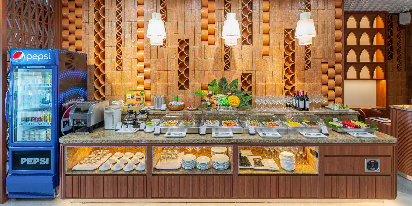 Buffet counter at SH Premium Lounge, Cam Ranh International Airport Terminal 1, offering a selection of hot dishes, fresh fruits, snacks, wine, and soft drinks in a modern wood-accented setting
