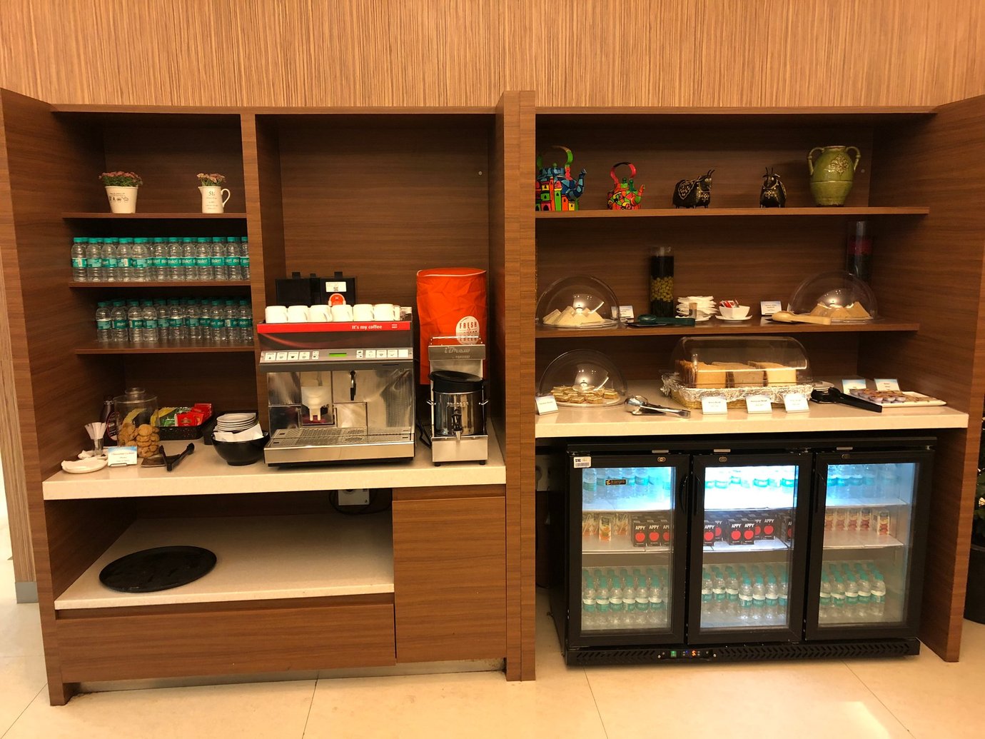 Self-serve buffet and beverage station at TFS Travel Club Lounge, Chennai Airport Terminal 4, featuring coffee machines, snacks, bottled water, refrigerated drinks, and neatly arranged food items.