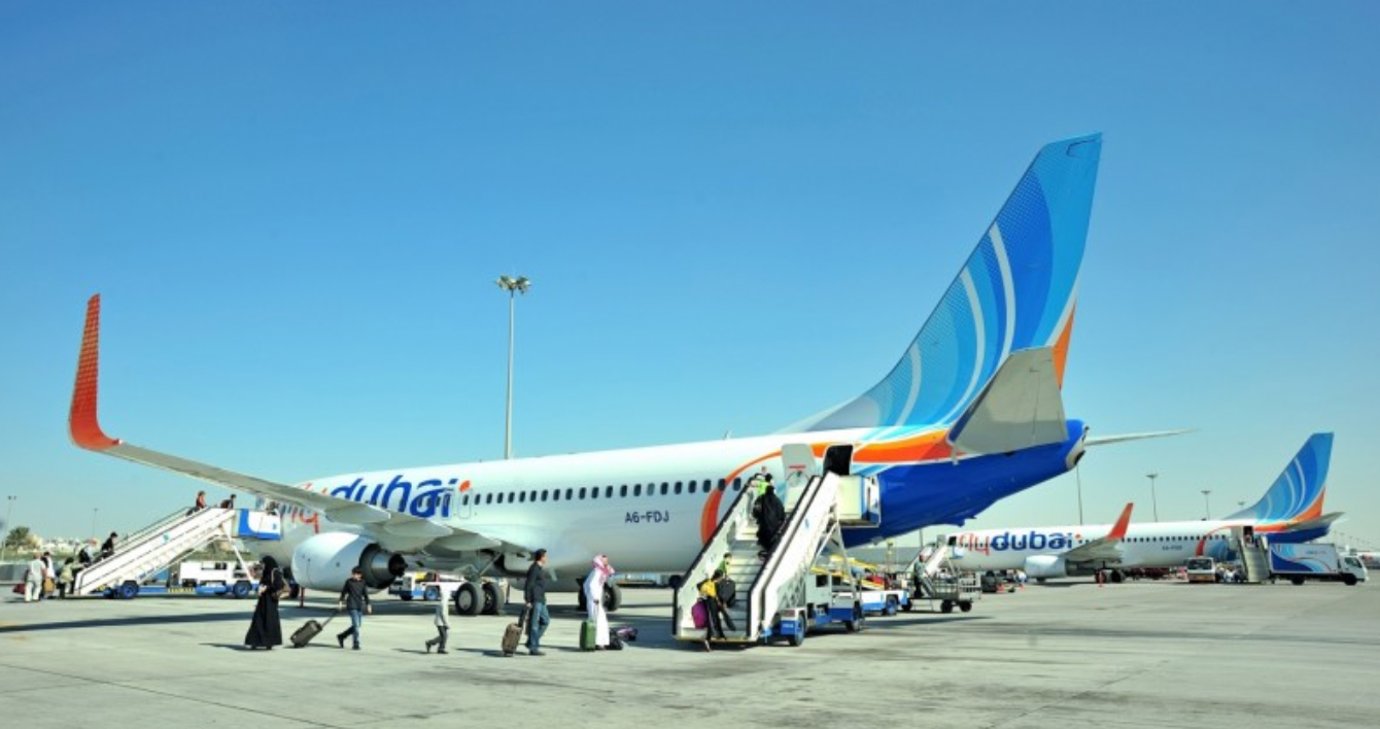 Passengers board a Flydubai aircraft via mobile stairs on the tarmac under a clear blue sky at Dubai International Airport. Several Flydubai planes with blue and orange tail designs are parked in the background, with ground staff and vehicles servicing the aircraft.