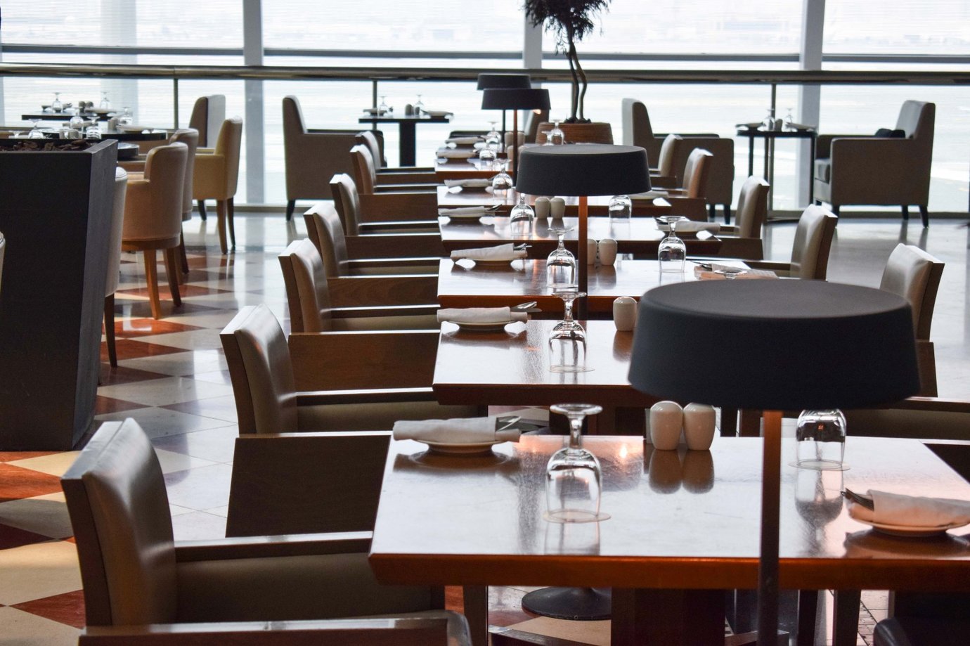 Dining area inside the Ahlan Lounge at Dubai International Airport Terminal 3 Concourse C, featuring neatly arranged wooden tables with glassware, modern black lamps, and panoramic runway views through floor-to-ceiling windows. Dining area inside the Ahlan Lounge at Dubai International Airport Terminal 3 Concourse C, featuring neatly arranged wooden tables with glassware, modern black lamps, and panoramic runway views through floor-to-ceiling windows.