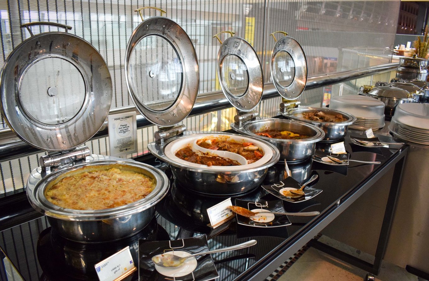 Buffet selection inside the Ahlan Lounge at Dubai International Airport Terminal 3 Concourse C, featuring a variety of hot dishes in stainless steel serving trays, offering international cuisine in a modern dining setup. Buffet selection inside the Ahlan Lounge at Dubai International Airport Terminal 3 Concourse C, featuring a variety of hot dishes in stainless steel serving trays, offering international cuisine in a modern dining setup.