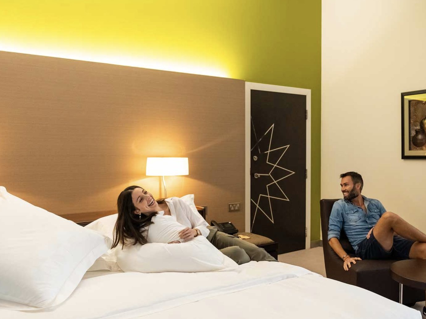 A couple relaxes in a bright, modern hotel room. A woman reclines on a large white bed laughing, while a man sits comfortably in a chair nearby, smiling at her. The room features warm lighting, minimalist decor, and a pop of lime green on the accent wall. A couple relaxes in a bright, modern hotel room. A woman reclines on a large white bed laughing, while a man sits comfortably in a chair nearby, smiling at her. The room features warm lighting, minimalist decor, and a pop of lime green on the accent wall.