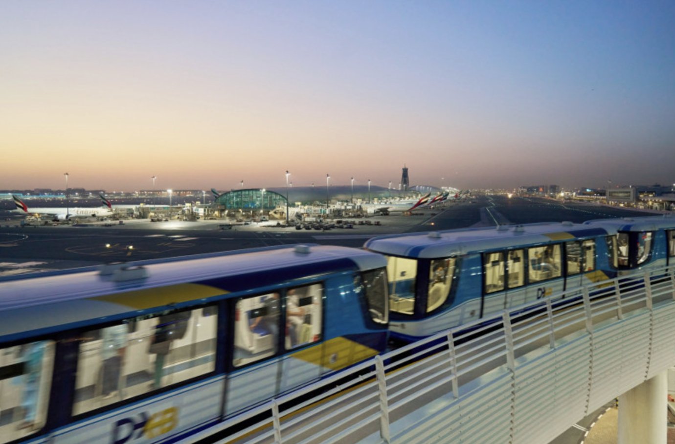 The Dubai International Airport (DXB) automated train glides along an elevated track at dusk, connecting terminals across the vast airfield. In the background, Emirates aircraft are parked at brightly lit gates beside the modern terminal buildings. The Dubai International Airport (DXB) automated train glides along an elevated track at dusk, connecting terminals across the vast airfield. In the background, Emirates aircraft are parked at brightly lit gates beside the modern terminal buildings.