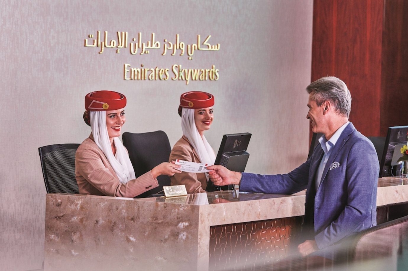 A man hands his boarding pass to an Emirates lounge agent at the Emirates Skywards reception desk. Two Emirates staff members, dressed in the airline’s signature beige uniforms and red hats, greet him with welcoming smiles. A man hands his boarding pass to an Emirates lounge agent at the Emirates Skywards reception desk. Two Emirates staff members, dressed in the airline’s signature beige uniforms and red hats, greet him with welcoming smiles.