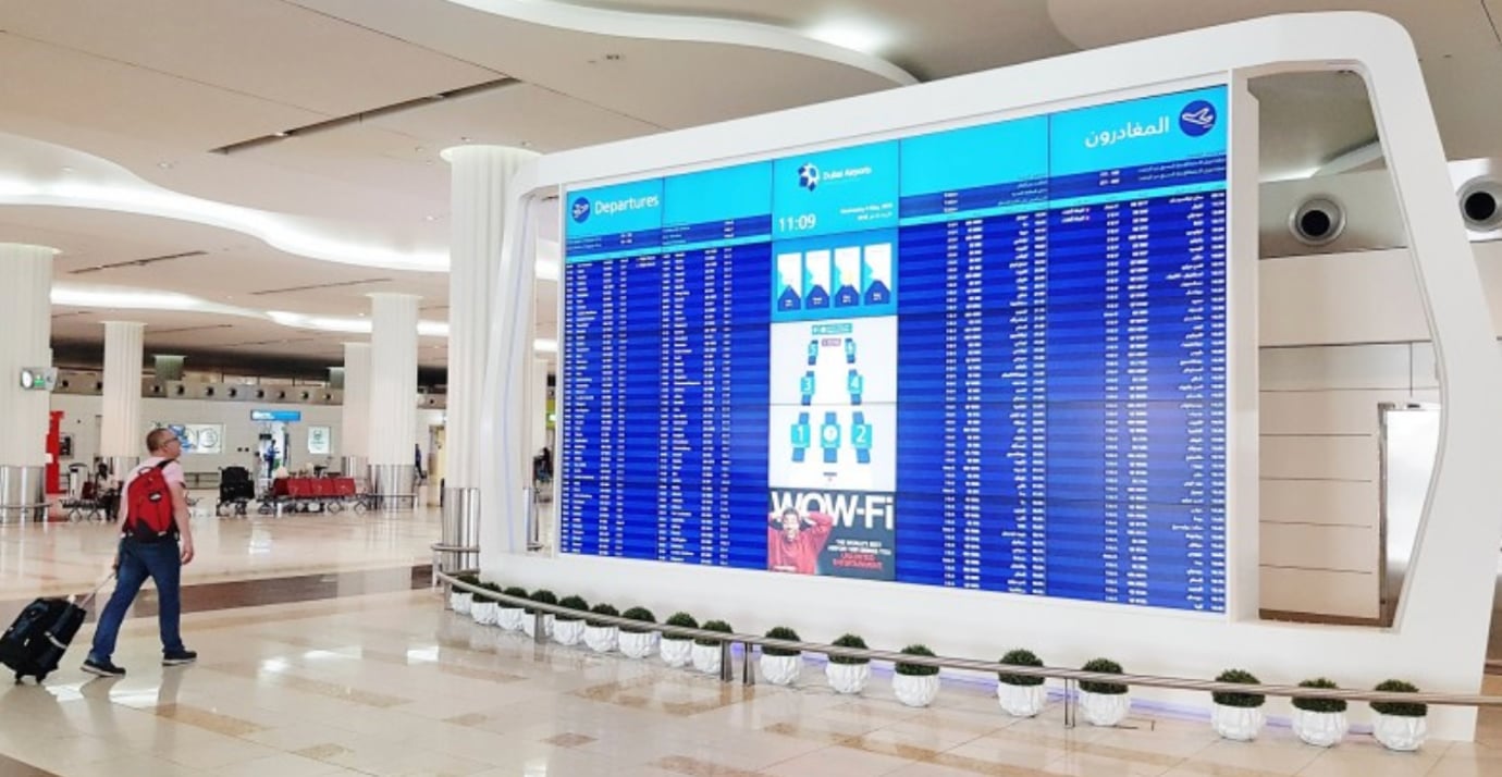 Large digital departures board in Dubai International Airport Terminal 3 showing flight information, with a traveler pulling luggage through the modern, bright concourse Large digital departures board in Dubai International Airport Terminal 3 showing flight information, with a traveler pulling luggage through the modern, bright concourse