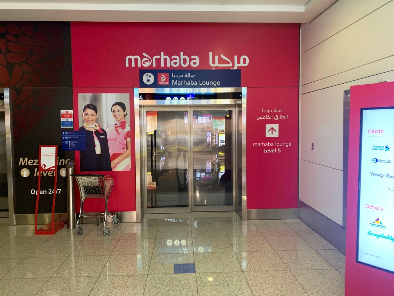 Entrance to the Marhaba Lounge at Dubai International Airport Terminal 3 Concourse A, featuring bright red signage, elevator access to Level 5, and promotional images of lounge staff welcoming guests. Entrance to the Marhaba Lounge at Dubai International Airport Terminal 3 Concourse A, featuring bright red signage, elevator access to Level 5, and promotional images of lounge staff welcoming guests.