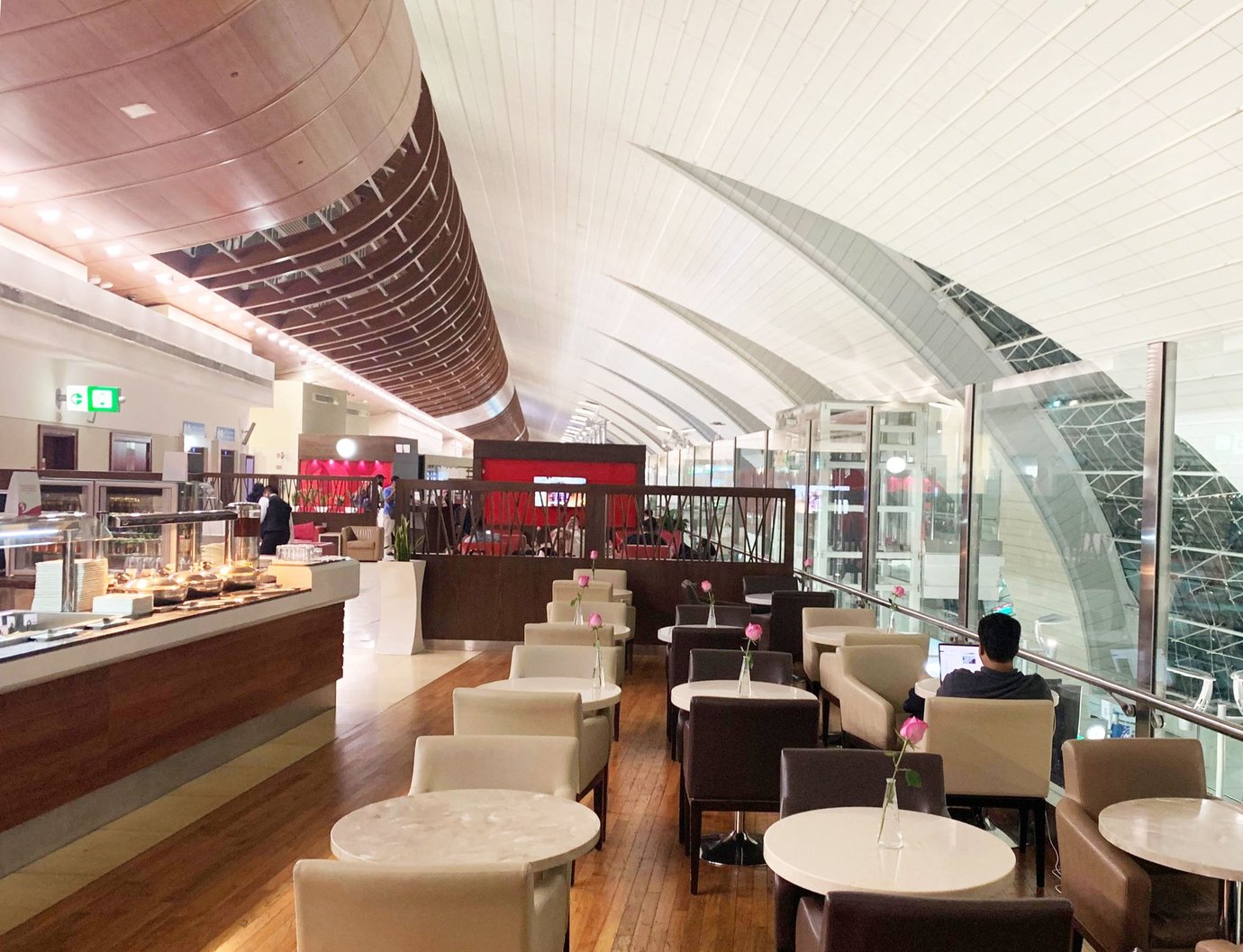 Dining and seating area inside the Marhaba Lounge at Dubai International Airport Terminal 3 Concourse B, featuring marble tables with single rose vases, leather chairs, buffet counters, and panoramic views of the terminal’s curved glass architecture. Dining and seating area inside the Marhaba Lounge at Dubai International Airport Terminal 3 Concourse B, featuring marble tables with single rose vases, leather chairs, buffet counters, and panoramic views of the terminal’s curved glass architecture.