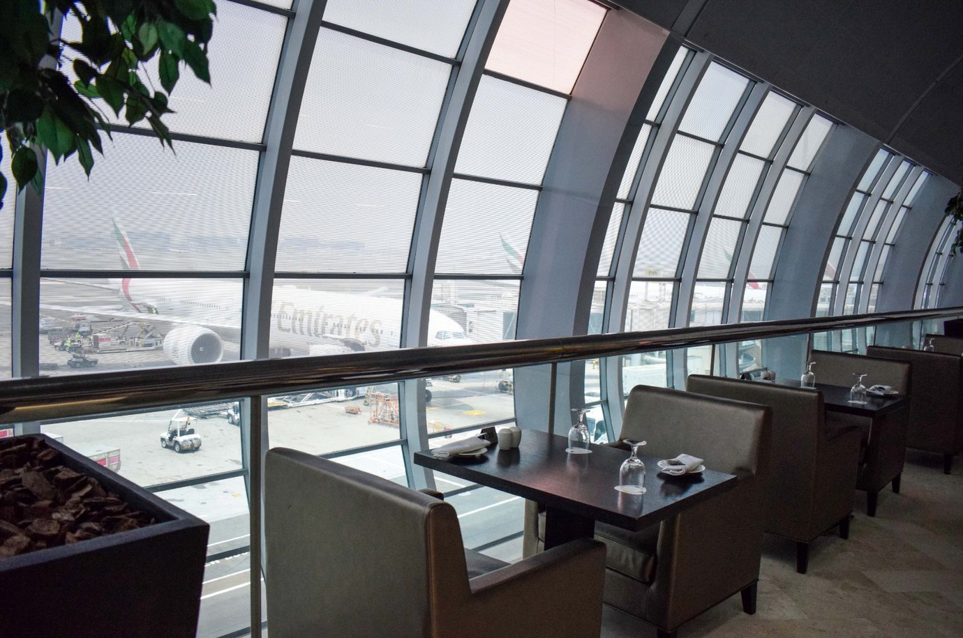 Dining tables overlooking the runway inside the Ahlan Lounge at Dubai International Airport Terminal 3 Concourse C, featuring elegant seating with glassware and panoramic views of Emirates aircraft through the curved glass windows Dining tables overlooking the runway inside the Ahlan Lounge at Dubai International Airport Terminal 3 Concourse C, featuring elegant seating with glassware and panoramic views of Emirates aircraft through the curved glass windows