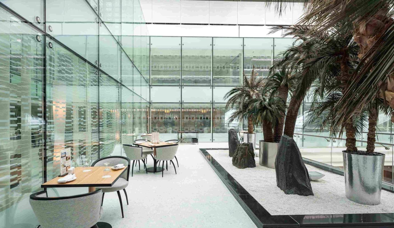 Modern seating area inside the Ahlan Lounge at Dubai International Airport Terminal 3 Concourse A, featuring glass walls, indoor palm trees, and elegant dining tables in a bright, tranquil setting Modern seating area inside the Ahlan Lounge at Dubai International Airport Terminal 3 Concourse A, featuring glass walls, indoor palm trees, and elegant dining tables in a bright, tranquil setting