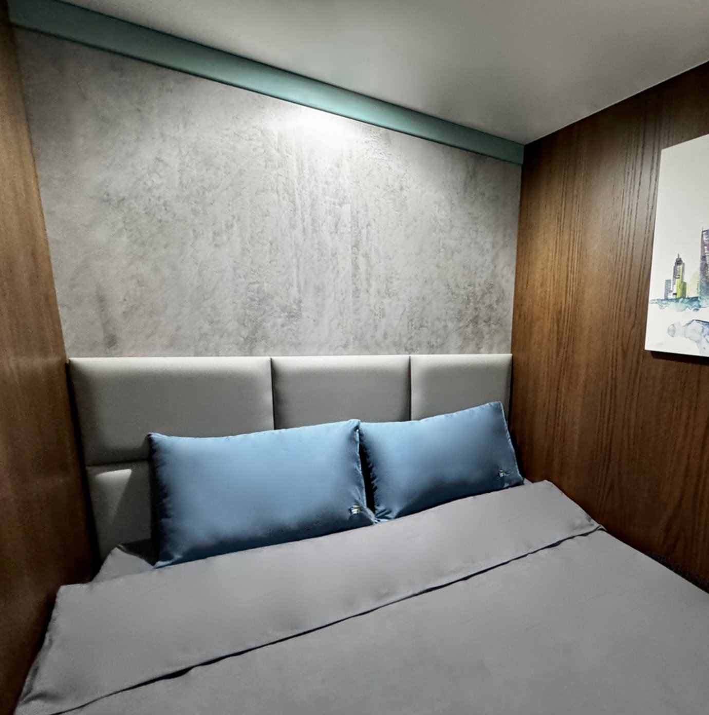 A compact sleep pod suite featuring a neatly made double bed with grey sheets and two blue satin pillows. The room has soft lighting, wood-paneled walls, and a minimalist grey upholstered headboard. A compact sleep pod suite featuring a neatly made double bed with grey sheets and two blue satin pillows. The room has soft lighting, wood-paneled walls, and a minimalist grey upholstered headboard.
