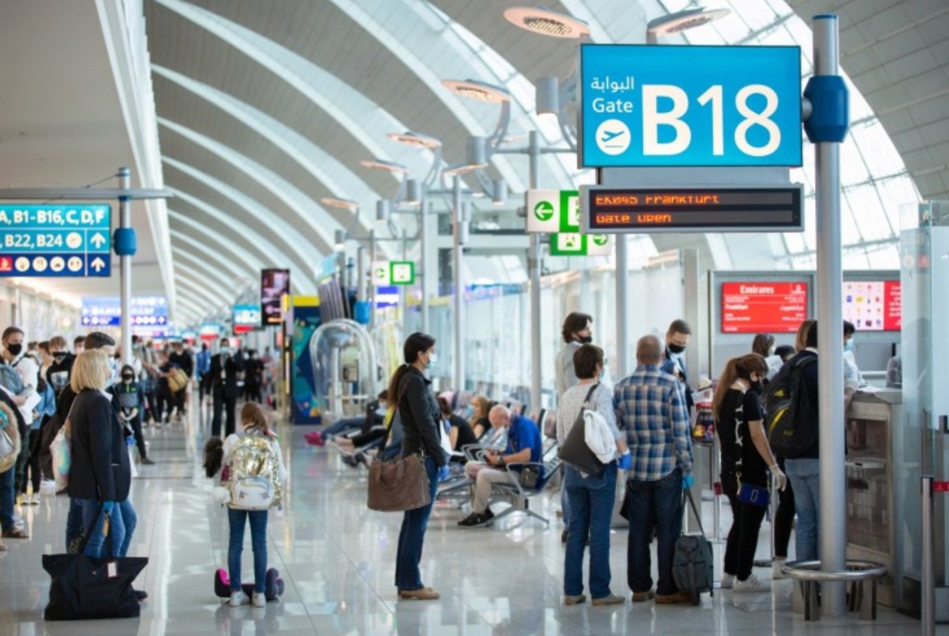 Passengers wait at Gate B18 in Dubai International Airport’s Terminal 3 concourse. The scene shows travelers standing in line and seated nearby, surrounded by clear directional signs and bright natural light filtering through the arched glass ceiling. Passengers wait at Gate B18 in Dubai International Airport’s Terminal 3 concourse. The scene shows travelers standing in line and seated nearby, surrounded by clear directional signs and bright natural light filtering through the arched glass ceiling.