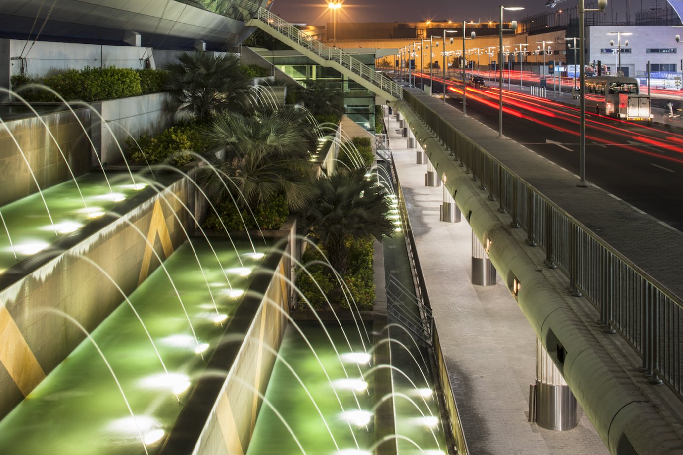 Dubai Airport exterior at night showing lit water fountains, greenery, and passing traffic near the terminal. Dubai Airport exterior at night showing lit water fountains, greenery, and passing traffic near the terminal.