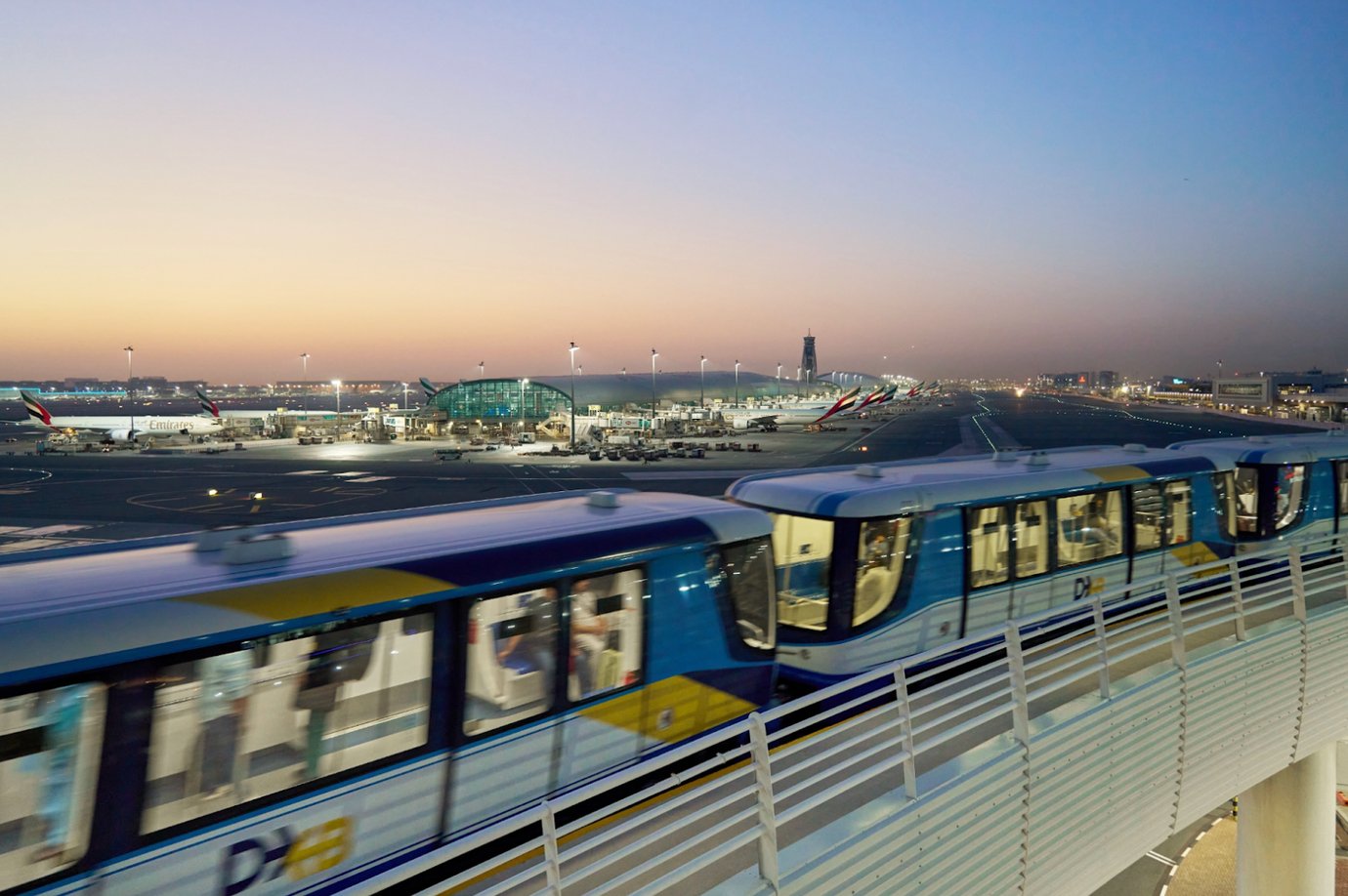 Dubai International Airport at dusk with the DXB automated train moving between terminals, Emirates aircraft parked at the gates, and the control tower visible in the background. Dubai International Airport at dusk with the DXB automated train moving between terminals, Emirates aircraft parked at the gates, and the control tower visible in the background.