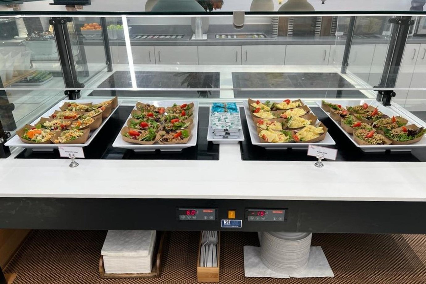 A food display at the Primeclass Lounge in JFK's Terminal 4, offering a selection of salads, sandwiches, and refreshments. The clean, modern buffet setup is well-maintained, with trays of food neatly presented under a glass cover and utensils. A food display at the Primeclass Lounge in JFK's Terminal 4, offering a selection of salads, sandwiches, and refreshments. The clean, modern buffet setup is well-maintained, with trays of food neatly presented under a glass cover and utensils.