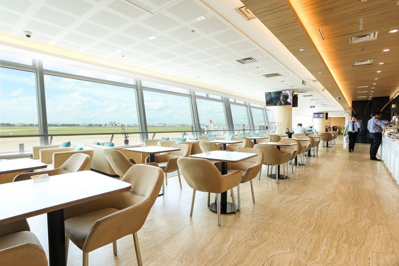 Bright seating area inside the Apricot Business Lounge at Tan Son Nhat Airport, with rows of beige upholstered chairs and dining tables set along floor-to-ceiling windows overlooking the runway. Bright seating area inside the Apricot Business Lounge at Tan Son Nhat Airport, with rows of beige upholstered chairs and dining tables set along floor-to-ceiling windows overlooking the runway.