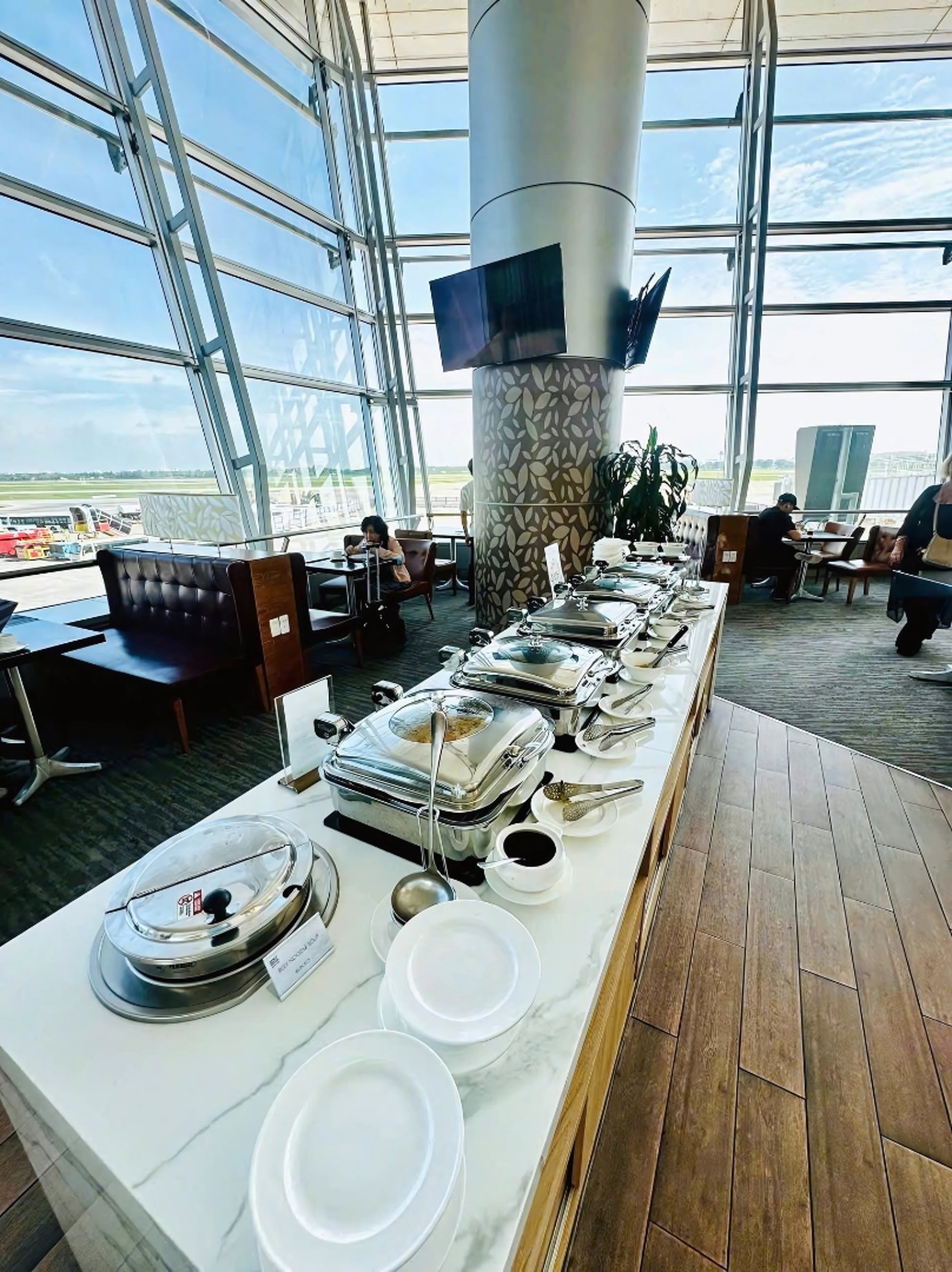 Sunlit airport lounge buffet with stainless chafing dishes on a marble counter, wood flooring, and runway views through floor-to-ceiling glass. Sunlit airport lounge buffet with stainless chafing dishes on a marble counter, wood flooring, and runway views through floor-to-ceiling glass.