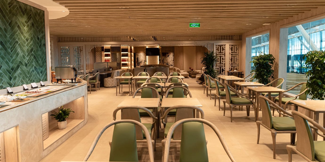 Wide view of the SH Premium Lounge dining area at Tan Son Nhat Airport, featuring a calm, wood-toned interior with slatted ceiling, green-upholstered chairs, neatly spaced dining tables, indoor plants, and a curved buffet counter along the left. Wide view of the SH Premium Lounge dining area at Tan Son Nhat Airport, featuring a calm, wood-toned interior with slatted ceiling, green-upholstered chairs, neatly spaced dining tables, indoor plants, and a curved buffet counter along the left.