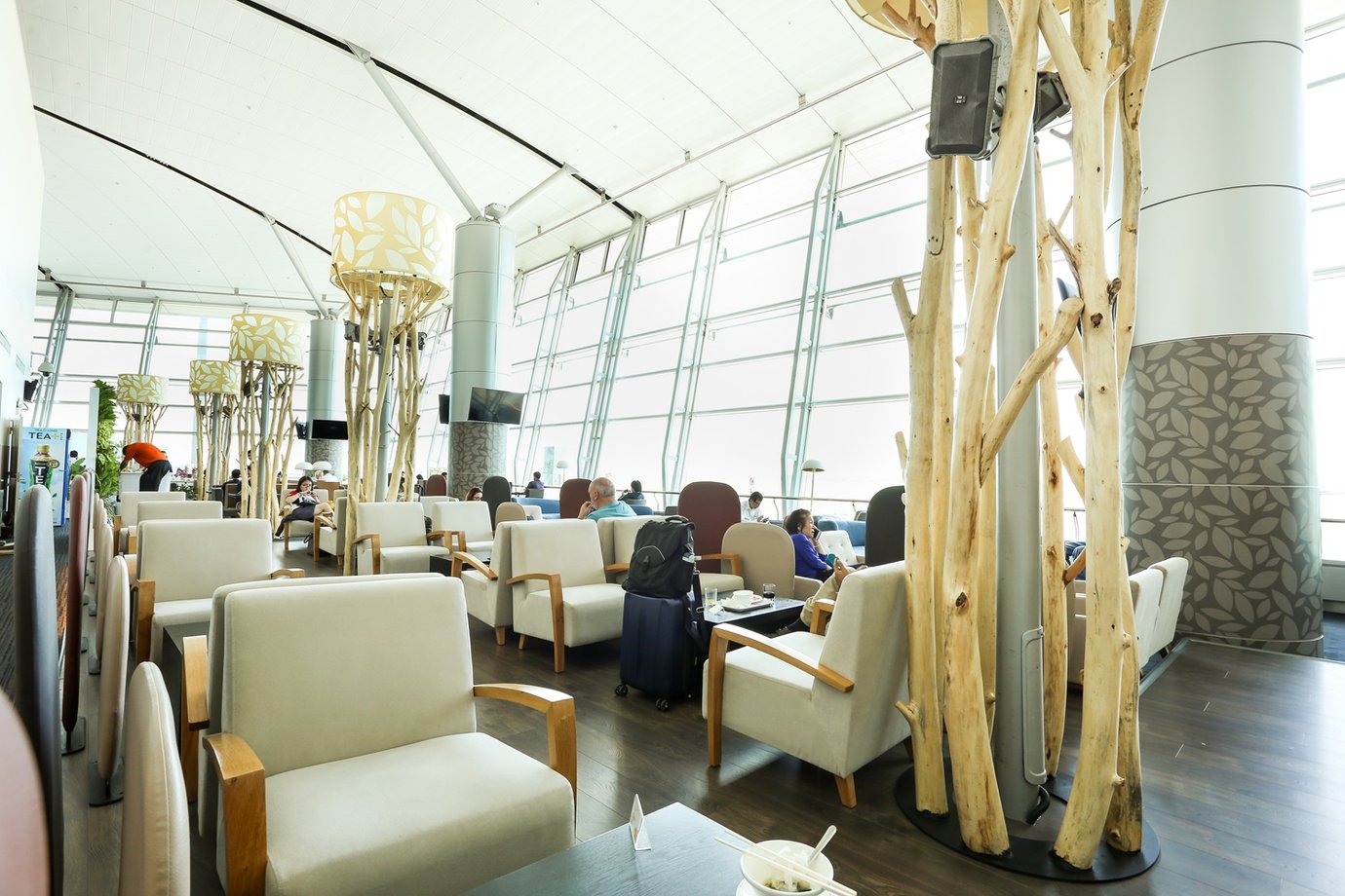 Bright airport lounge with cream armchairs, wood accents, sculptural tree-like columns, and large windows overlooking the runway. Bright airport lounge with cream armchairs, wood accents, sculptural tree-like columns, and large windows overlooking the runway.