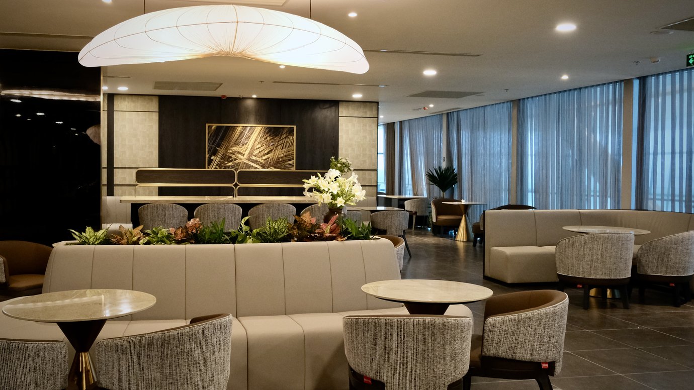 Seating area in The Sens Business Class Lounge at Tan Son Nhat Airport, showing curved upholstered sofas and fabric armchairs arranged around small marble-topped tables, with low planters of greenery. Seating area in The Sens Business Class Lounge at Tan Son Nhat Airport, showing curved upholstered sofas and fabric armchairs arranged around small marble-topped tables, with low planters of greenery.