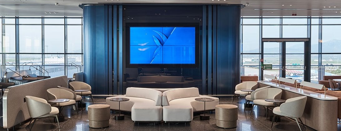 Media area in the Aegean business lounge at Athens airport. The space is centred around a large digital screen. The room is framed by large windows offering views of the airport and mountains in the distance. The seating is modern in neutral tones.