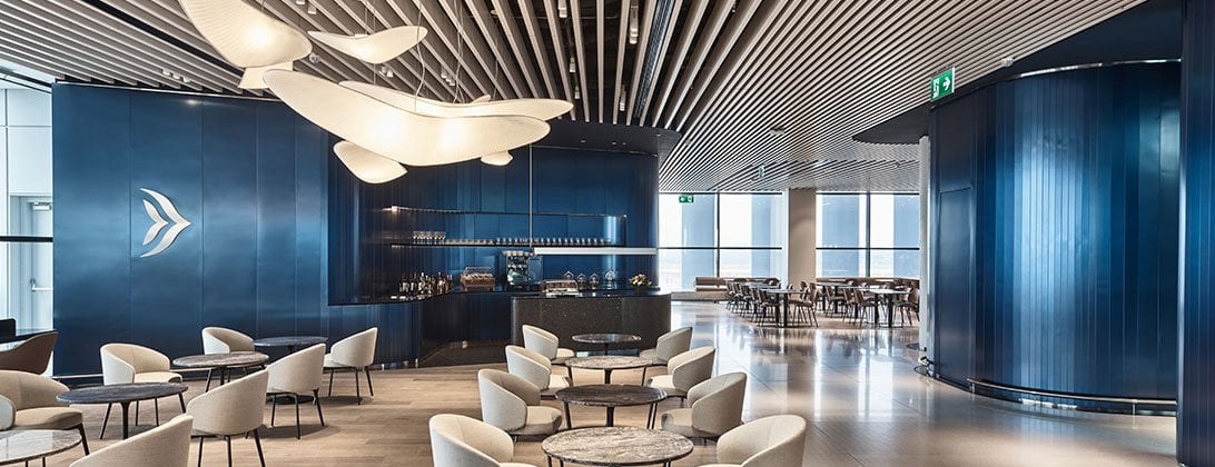A sophisticated bar and seating area inside the Aegan Business Lounge at Athens Airport, featuring a striking blue wall with the logo illuminated. The space is furnished with plus light coloured chairs, and marble tables.