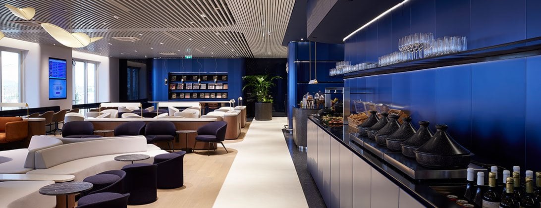 View of seating and buffet section of the Aegean Business Lounge in Hall B, Athens Airport. There is various seating to the left of the room. In the buffet food is covered in elegant cone shaped covers, and there is an assortment of alcoholic drinks.